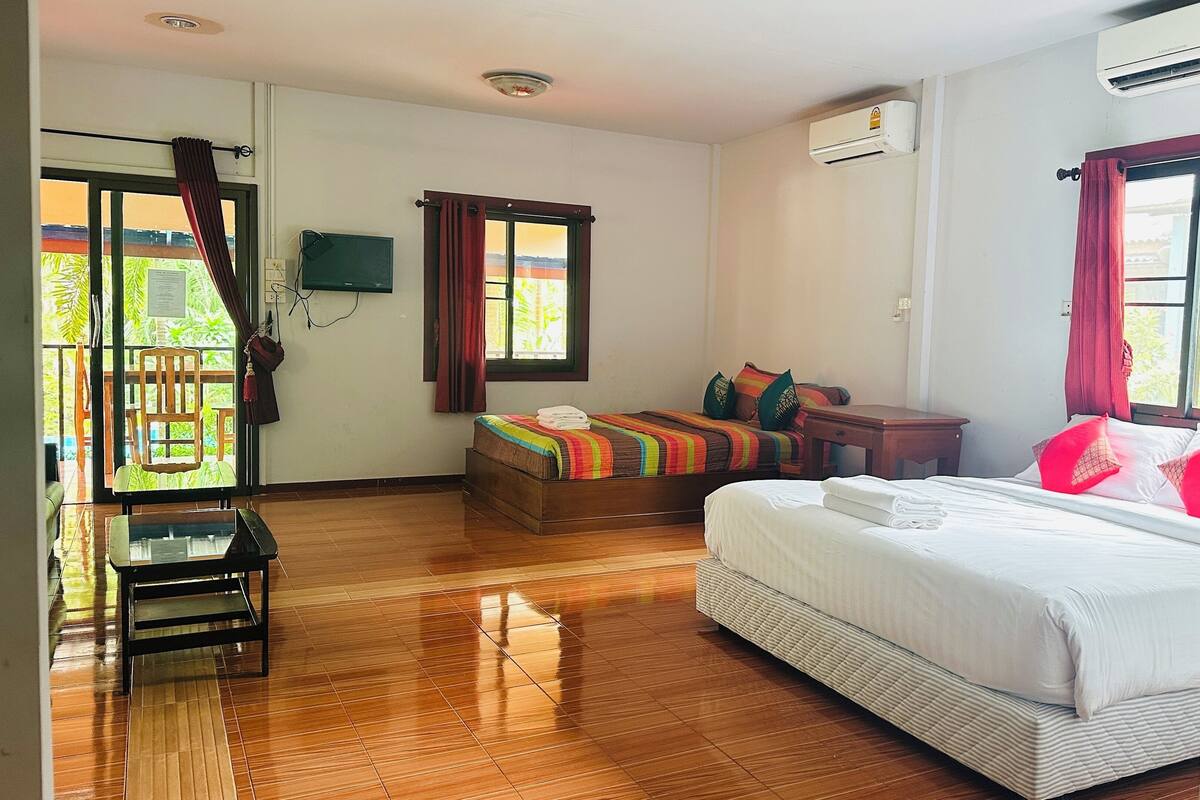 A hotel room with a wooden floor, a bed with white bedding, a wooden nightstand, a small table, and a chair. There is a window with curtains, an air conditioning unit, and a view of greenery outside.