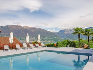 Piscina com espreguiçadeiras brancas e guarda-sóis, cercada por vegetação e montanhas ao fundo.