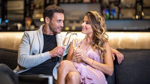 Een koppel zit op een bankje met champagneglazen, in een modern interieur met boekenplanken op de achtergrond.