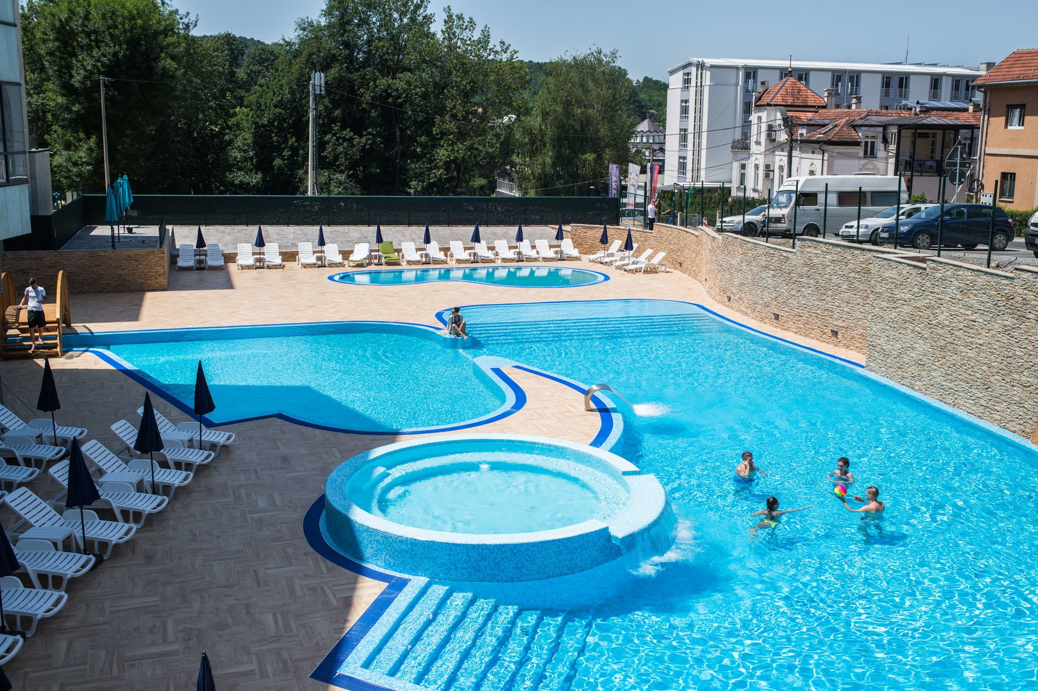 A swimming pool with lounge chairs and umbrellas, surrounded by a stone wall and buildings in the background.