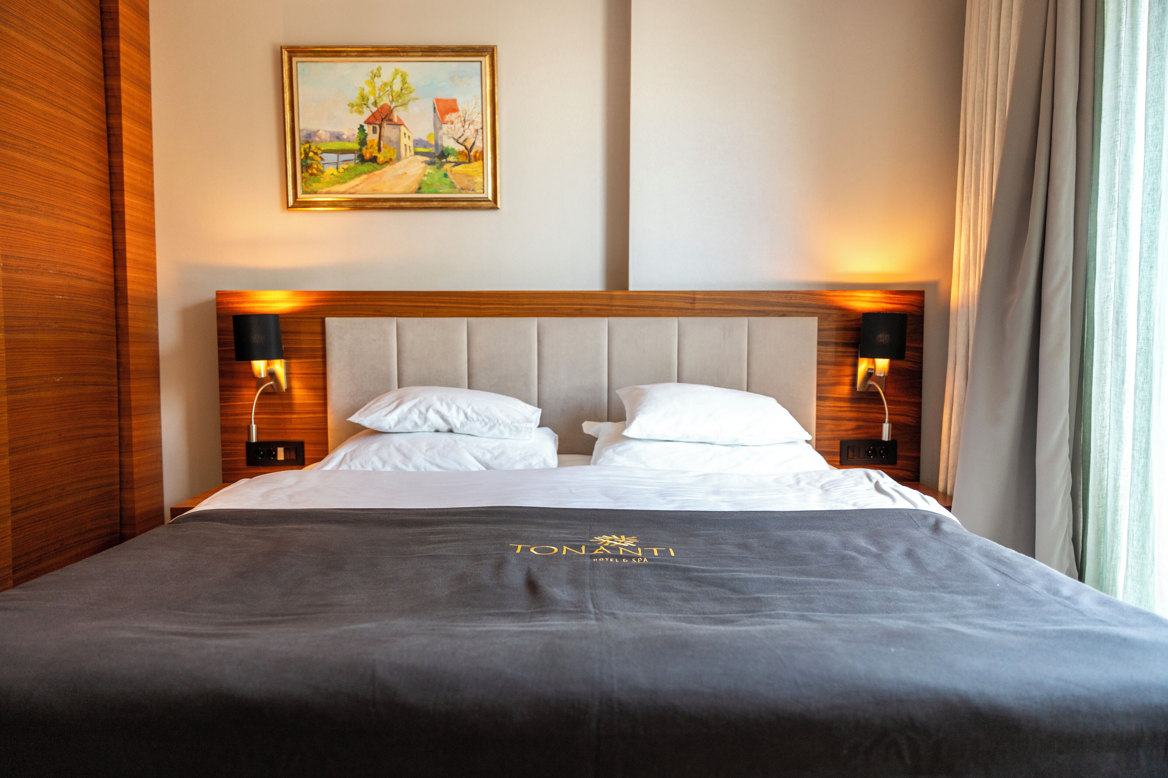 A hotel room with a bed, two bedside lamps, a painting on the wall, and a window with curtains.