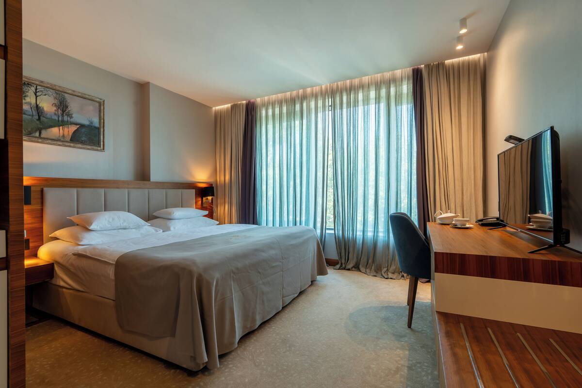 Habitación de hotel con cama, escritorio con computadora, silla y ventana grande con cortinas.