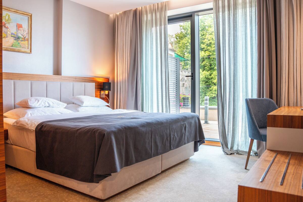 Habitación de hotel con cama, escritorio, silla, ventana con cortinas y un cuadro en la pared.
