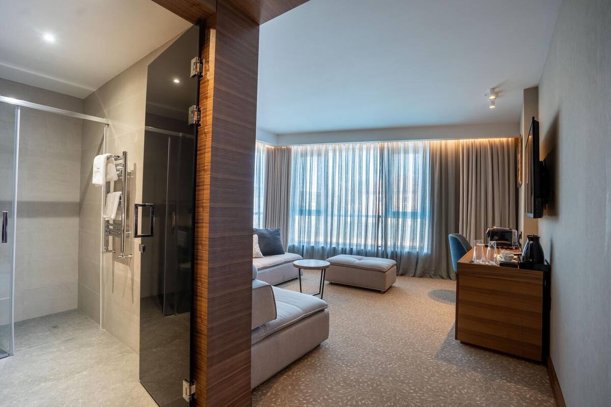 Una habitación de hotel moderna con ducha de vidrio, un sofá, una mesita redonda, un escritorio de madera y un televisor montado en la pared.