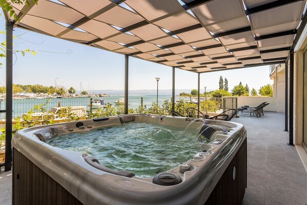 Relax in a serene hot tub under a shaded pergola, surrounded by lounge chairs and a well-maintained lawn with a marina view.