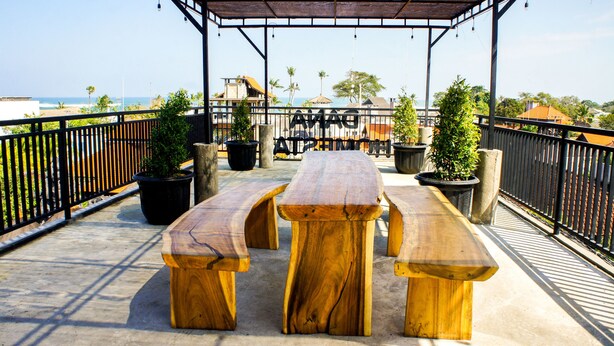 An outdoor wooden deck with picnic tables and potted plants, overlooking a beach and ocean. - Dana Homestay (Tibubeneng)