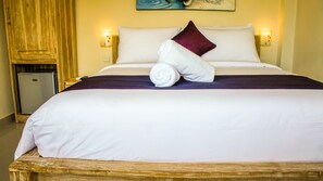 A neatly made bed with white linens and a purple accent pillow, a wooden headboard, and a small refrigerator in the corner.