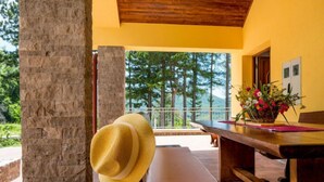 A wooden bench with a straw hat, a wooden table with a vase of flowers, and a view of trees and mountains through a balcony.