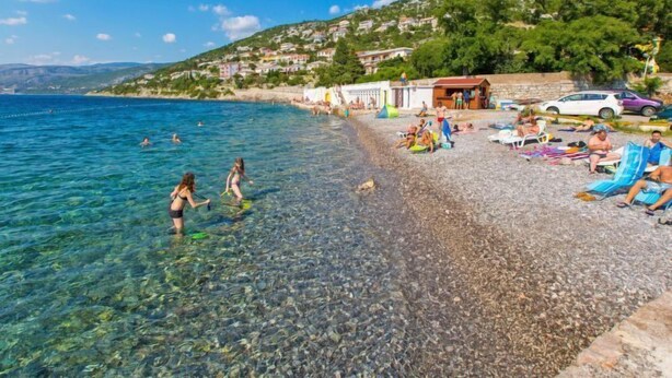 A beach with clear shallow waters, people swimming and sunbathing, and houses in the background.