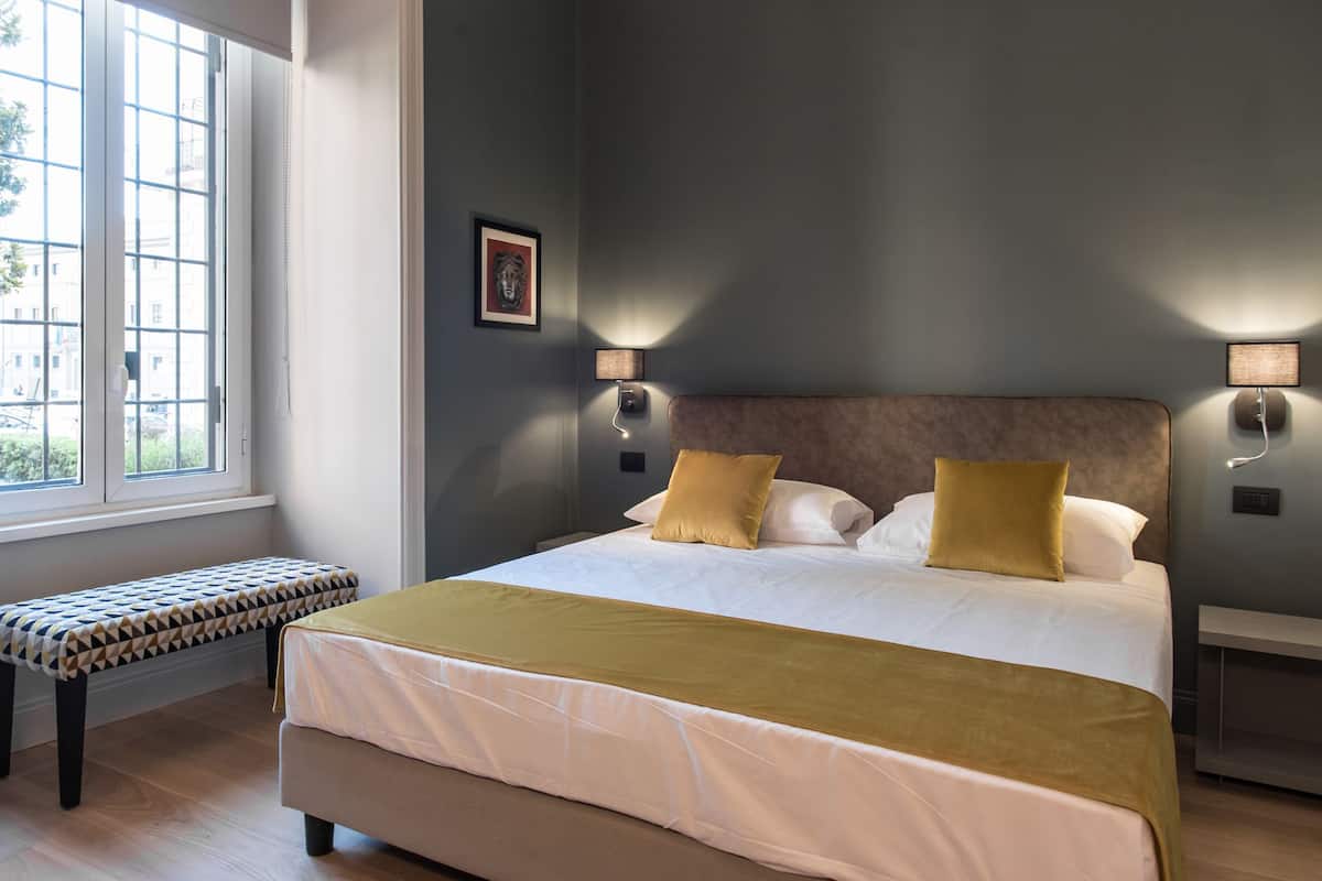 Una cama bien hecha con cabecera, dos lámparas de noche, un banco y una ventana con vistas a edificios.