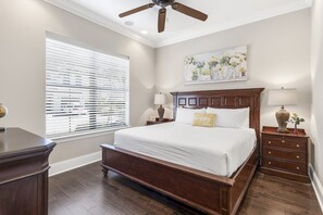 A bedroom with a large bed, wooden headboard, two bedside tables with lamps, a ceiling fan, and a window with blinds. - Pet Friendly ~ Two Golf Carts ~ Golf Course View (Miramar Beach)