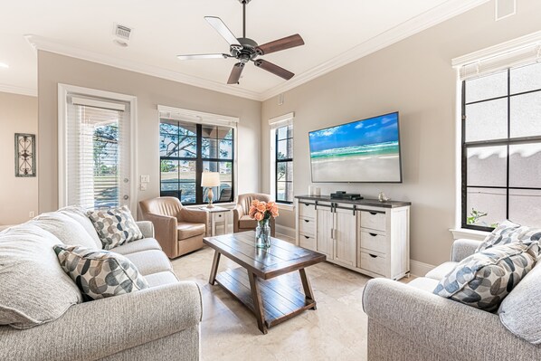 A living room with a sofa, coffee table, and TV. There are windows, a ceiling fan, and a kitchen area visible. - Pet Friendly ~ Two Golf Carts ~ Golf Course View (Miramar Beach)