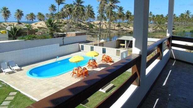 A pool area with lounge chairs and umbrellas, a view of the beach and palm trees, and a covered deck. - Pousada Solar da Lagoa Baixio (Esplanada)