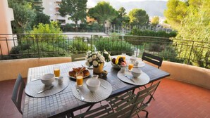 Un balcón con una mesa dispuesta para el desayuno, que incluye croissants, fruta y bebidas, con vistas a un paisaje verde con árboles y edificios.