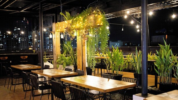 An outdoor dining area with wooden tables and black chairs, surrounded by potted plants and string lights.