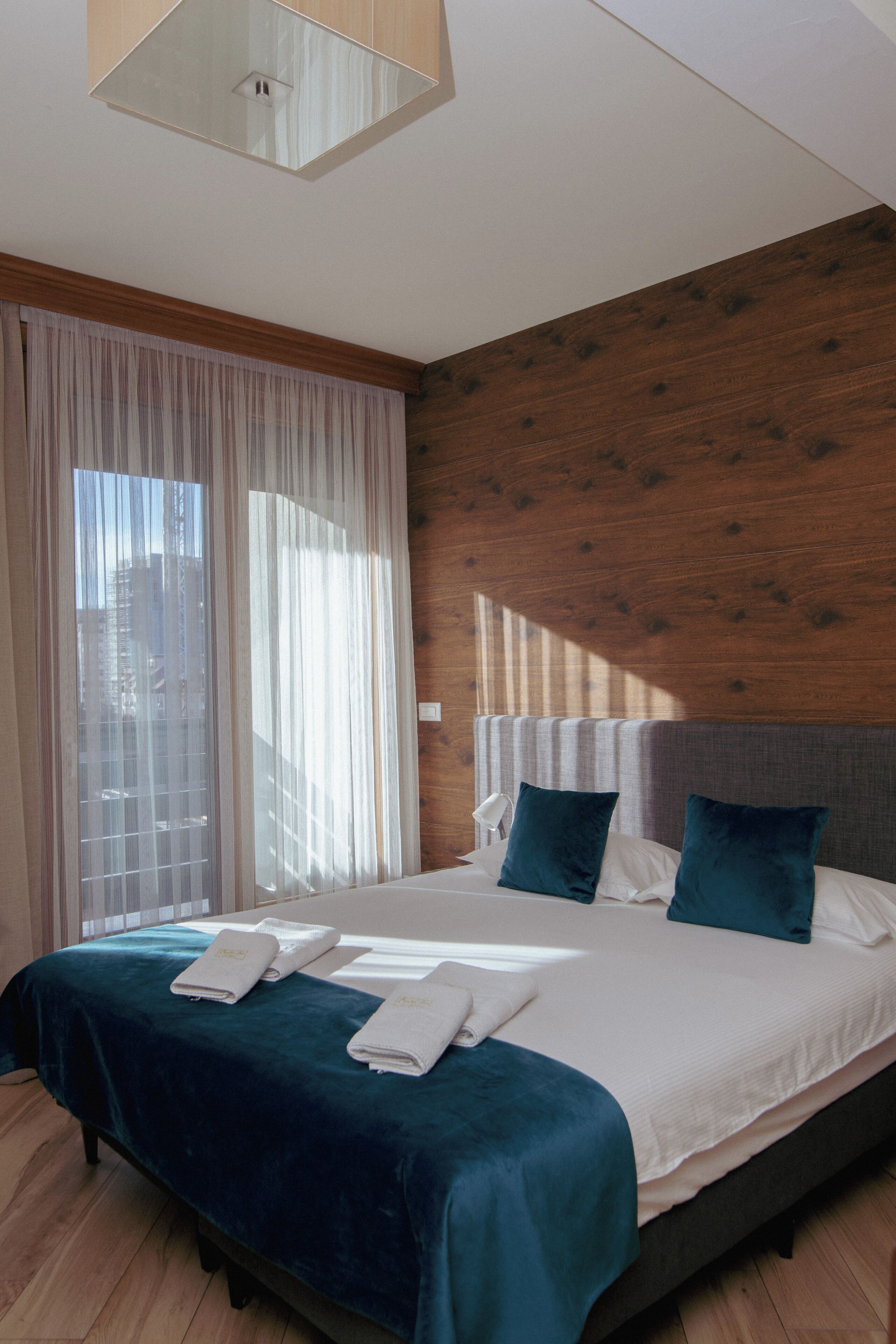 Quarto de hotel com cama, cortinas, parede de madeira e janela.
