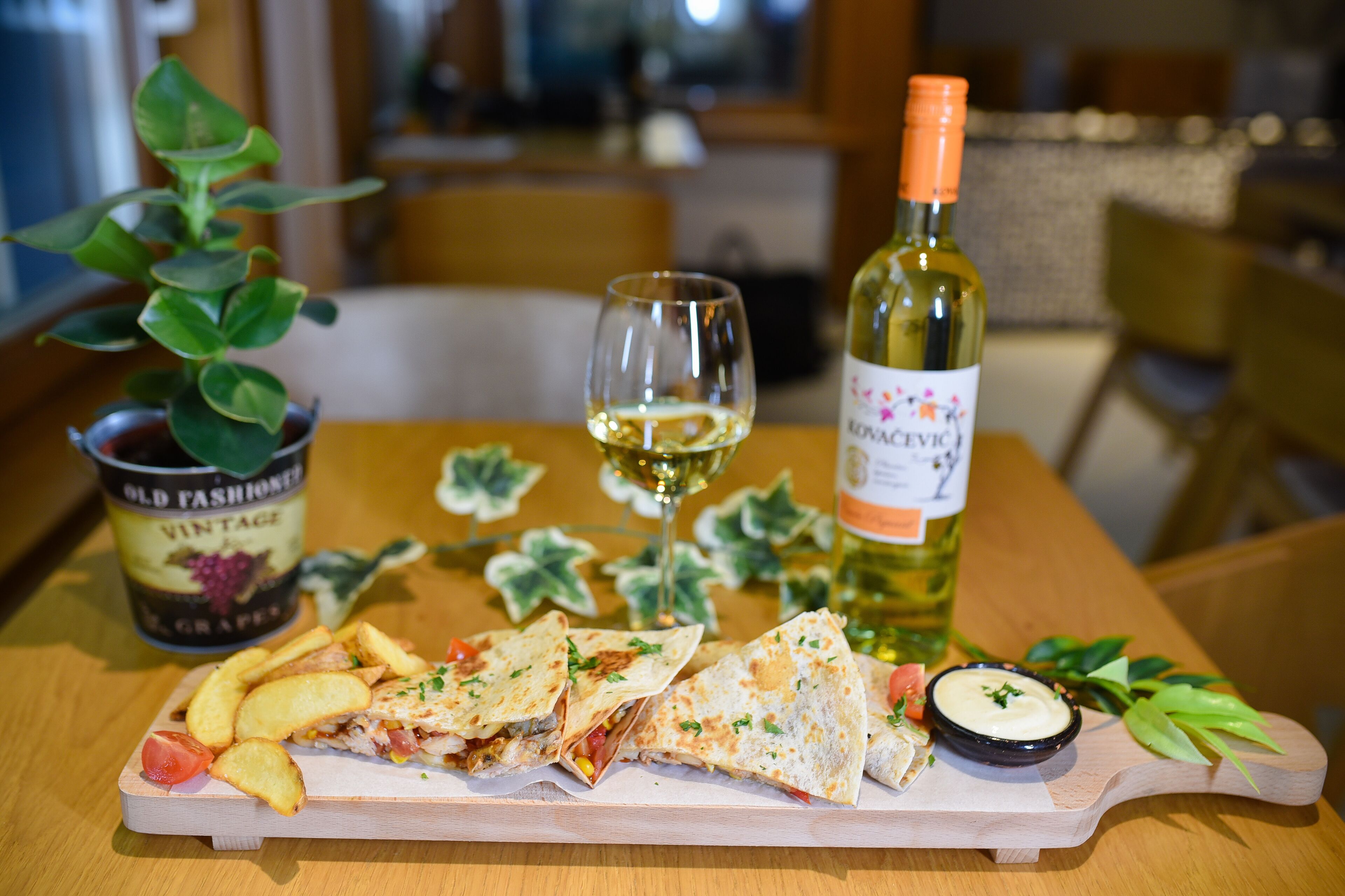 A wooden table with a meal consisting of quesadillas, chips, and a small bowl of sauce. A bottle of wine and a glass are also on the table.