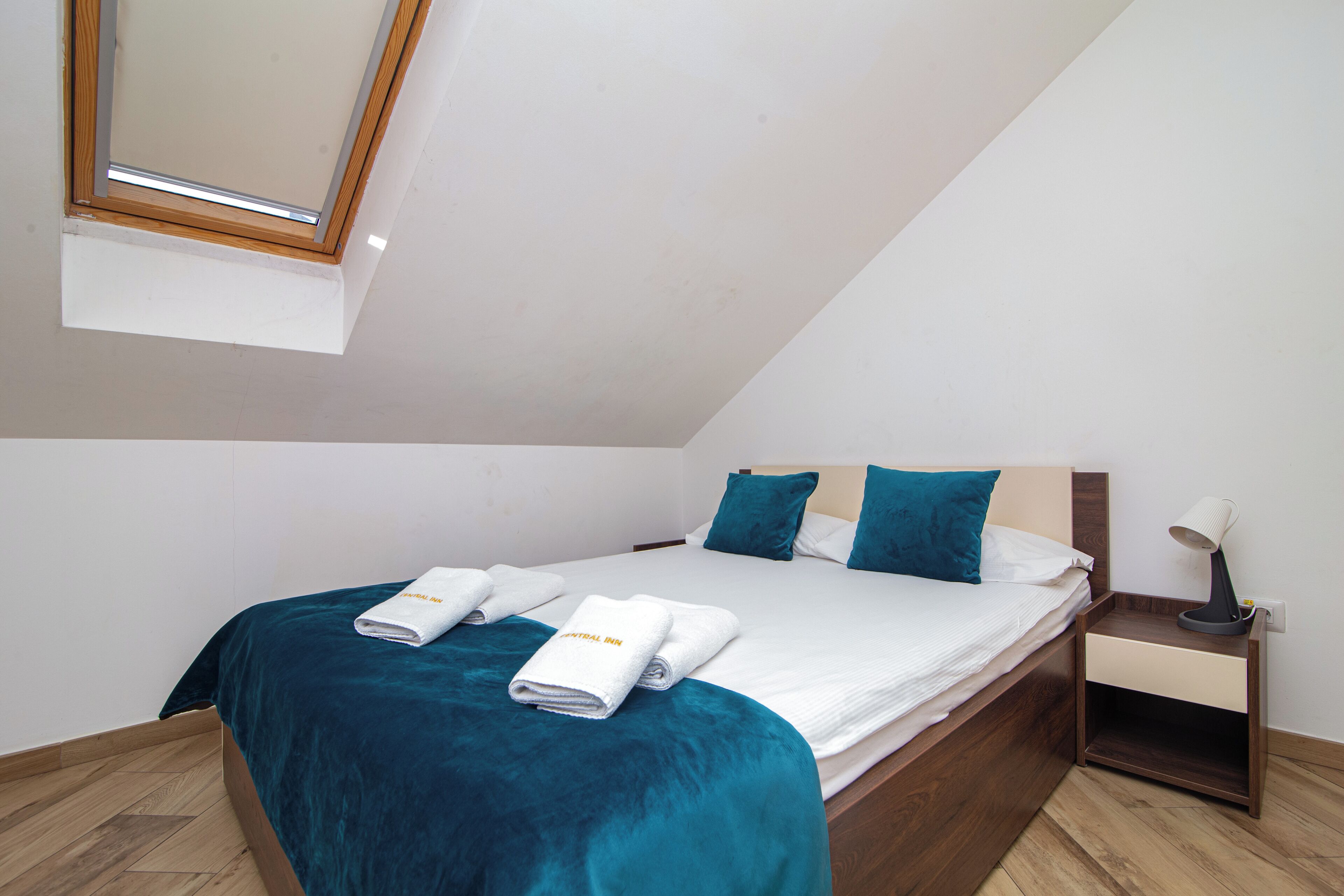 A bedroom with a skylight, a bed with teal bedding, a nightstand with a lamp, and a wooden headboard.