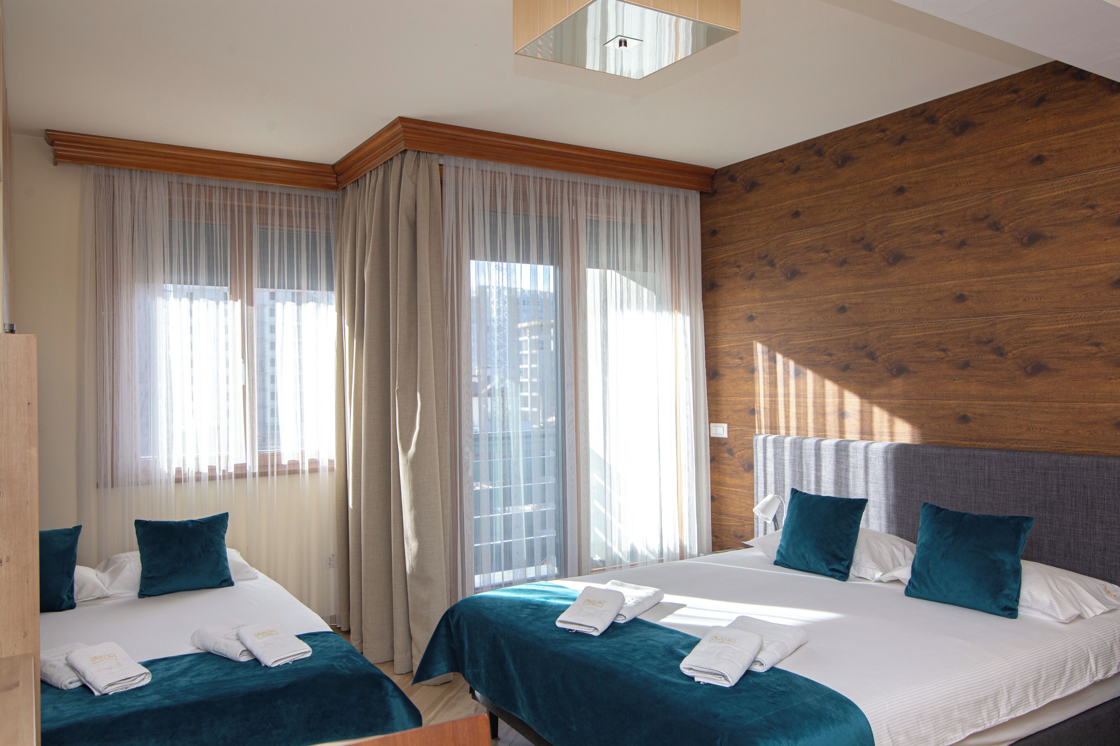 A hotel room with two beds, a wooden accent wall, and a large window with sheer curtains.