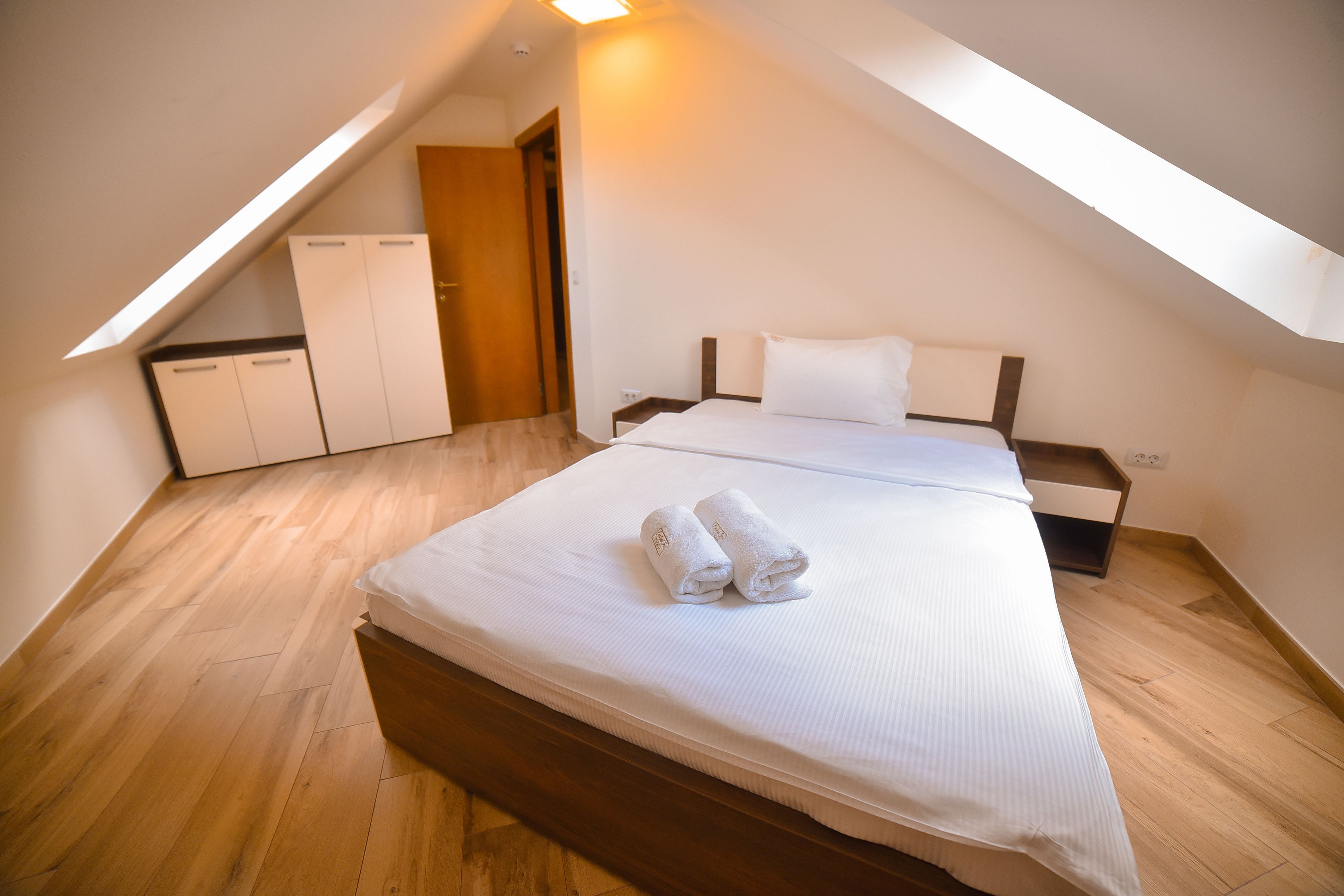 Un dormitorio con techo inclinado, suelo de madera, cama con ropa blanca y un armario.