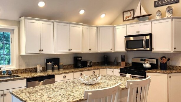 A modern kitchen with white cabinets, granite countertops, a central island with two white chairs, a skylight, and a sign that reads "Chatham." - Nautical Comfort near Rail Trail (Chatham)