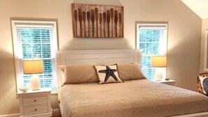 A bedroom with a large bed, two bedside tables with lamps, a decorative wall piece, and a view through the windows. - Nautical Comfort near Rail Trail (Chatham)