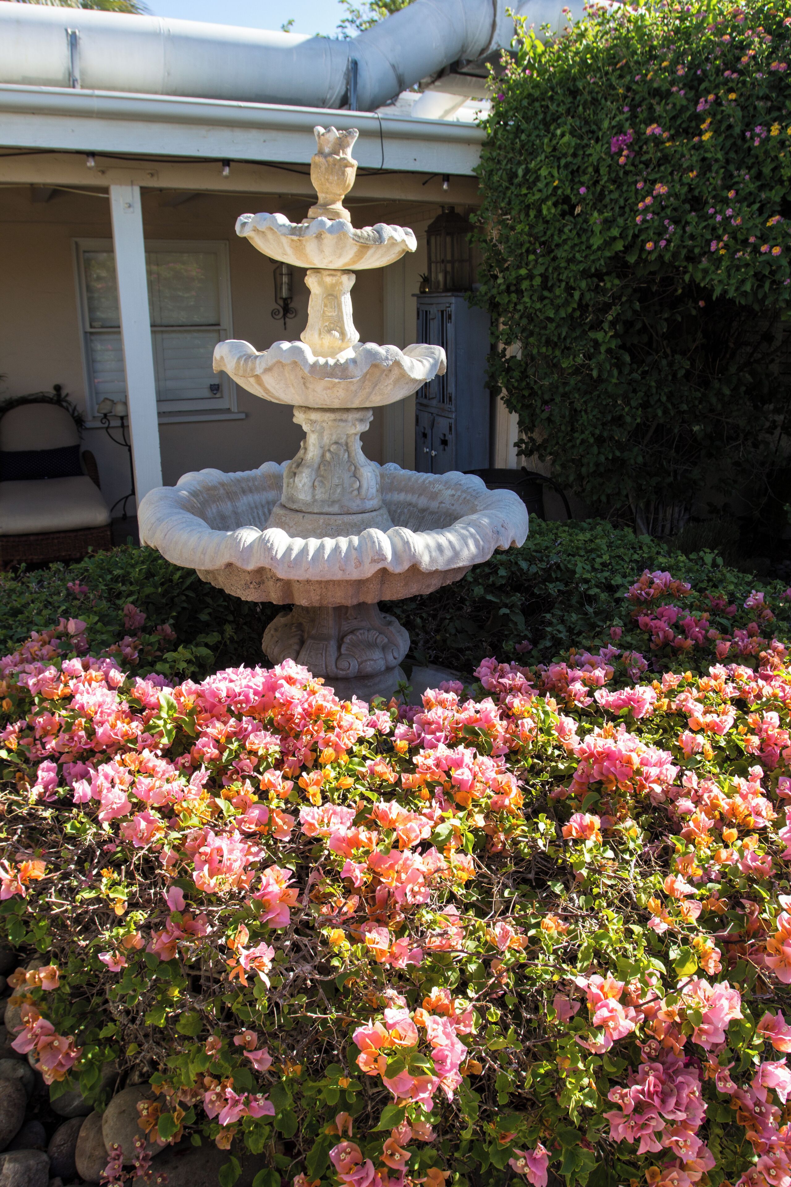 A multi-tiered stone fountain surrounded by pink flowers in a garden area.