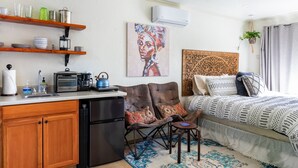Un espacio habitable pequeño con kitchenette, una cama con ropa de cama estampada, cafetera y un cuadro en la pared.