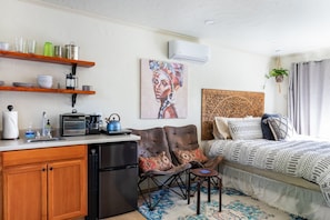 A compact living space with a kitchenette, a bed with patterned bedding, a coffee maker, and a painting on the wall.