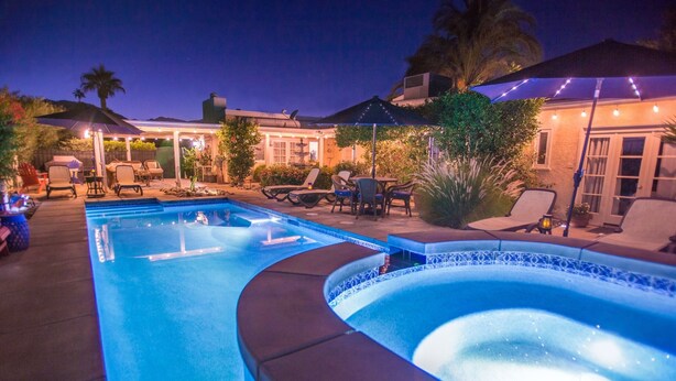 Una piscina con sillas de descanso y sombrillas, rodeada por un patio con plantas y un edificio al fondo.