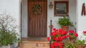 Un edificio de paredes blancas con una puerta de madera, una ventana y una maceta con flores rojas.