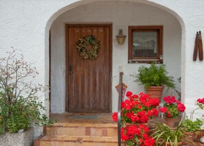 Un bâtiment aux murs blancs, doté d’une porte en bois, d’une fenêtre et d’un pot de fleurs contenant des fleurs rouges.