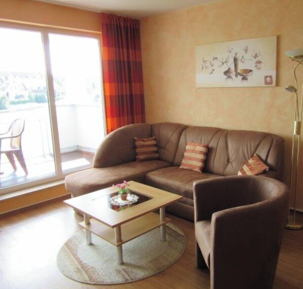 A living room with a brown sofa, a small coffee table with a vase, a chair, a painting on the wall, and a view through a sliding door. - (278) 2-room apartment - apartment complex Yachthafenresidenz (Kühlungsborn)