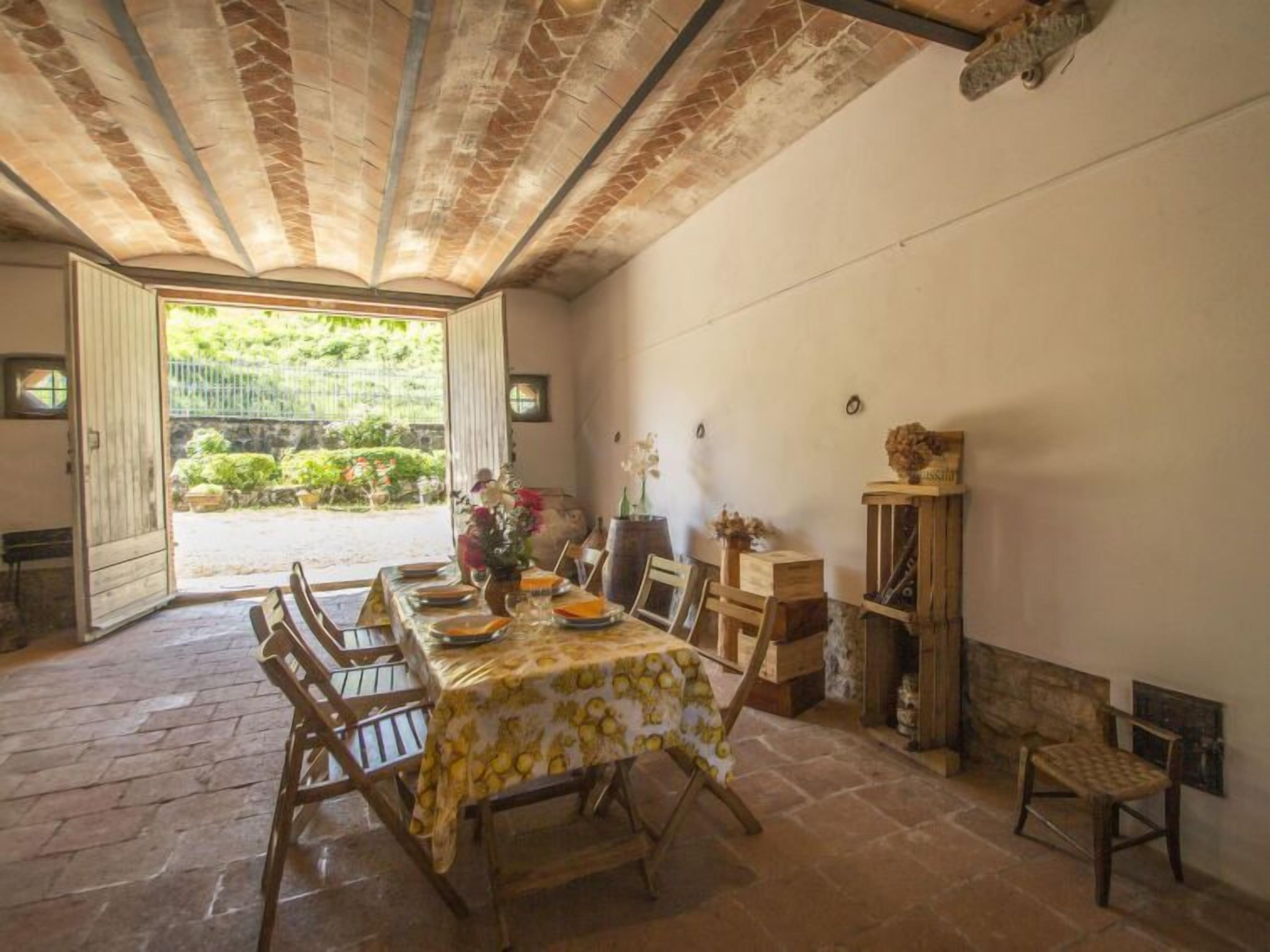 A dining area with a table set for four, surrounded by wooden chairs. The room features a stone floor, a high ceiling with exposed beams, and a view of a garden through an open door.