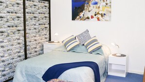 A bedroom with a brick-patterned wall, a bed with blue bedding, patterned pillows, a nightstand with a lamp, and a framed picture of a coastal village. - The Arches -Ultimate Upper Hunter Getaway- Large Entertainment Living&Relaxation (Singleton)