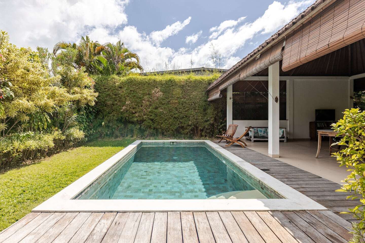 Een rechthoekig zwembad met een houten terras, omringd door weelderige beplanting en een overdekte patio met zitplaatsen.