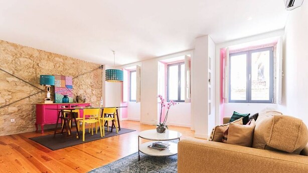 A living room with a wooden floor, a pink and yellow table, a beige sofa, and a patterned rug.