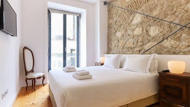 A bedroom with a large bed, a wooden floor, a stone wall, a window with a view, and a TV mounted on the wall.