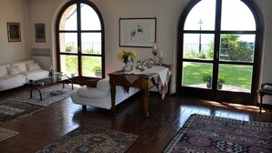 A spacious living room with a white sofa, a wooden coffee table, and a dining table set with flowers. Large arched windows provide views of a garden.
