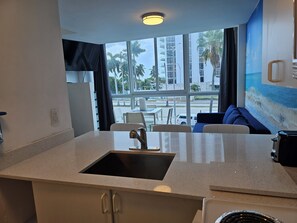 A modern kitchen with a sink, a dining area with chairs, and a large window offering a view of palm trees and buildings.