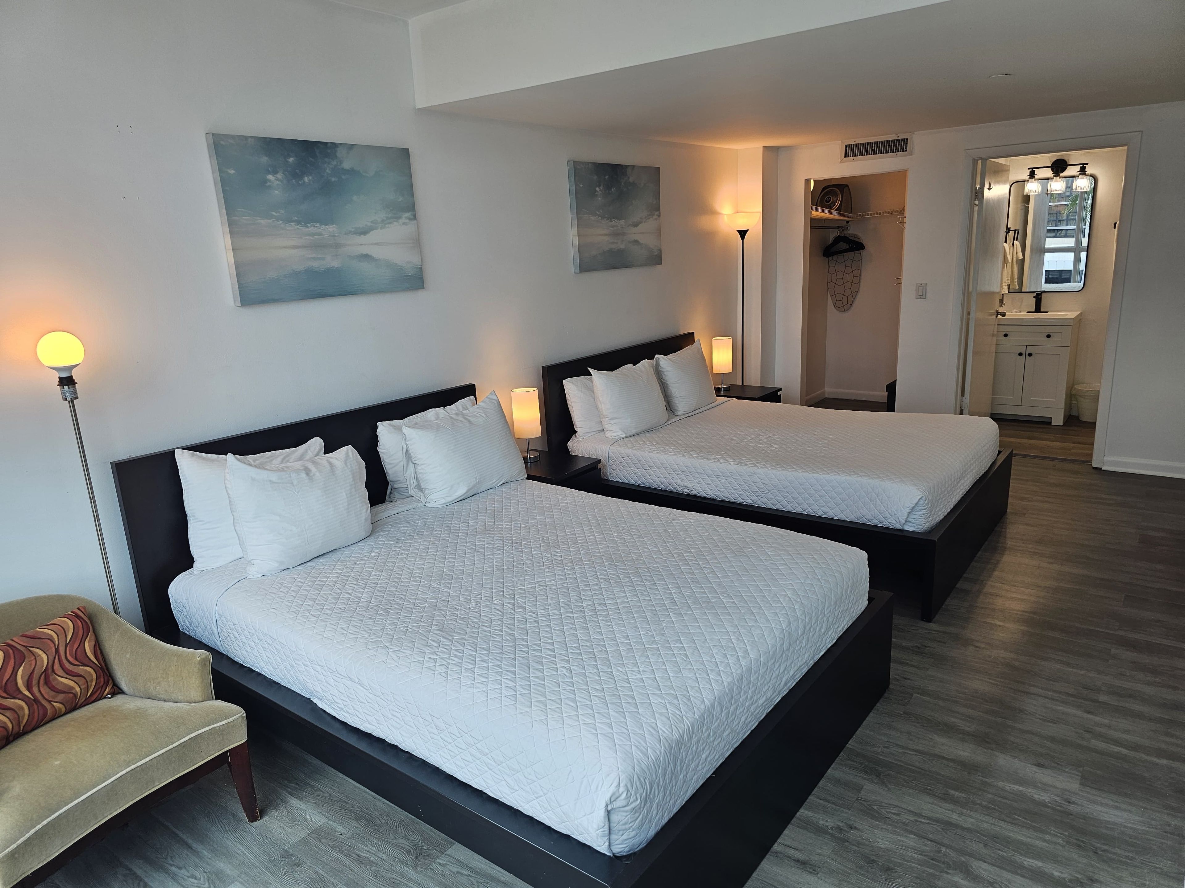 A hotel room with two beds, a chair, a lamp, and artwork on the walls.