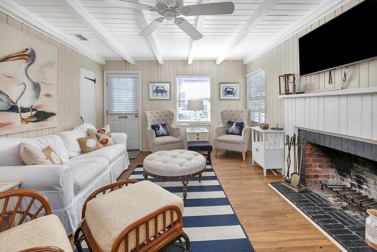 Uma sala de estar aconchegante com um sofá branco, duas poltronas, um banquinho redondo, uma lareira e um ventilador de teto.
