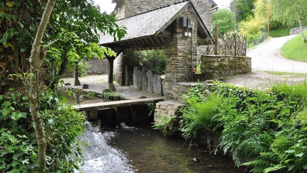 Ein Steinhaus mit einer Holzbrücke über einem Bach, umgeben von üppigem Grün.
