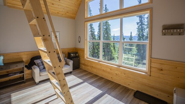Una habitación con techo inclinado, un ventanal con vistas a árboles, una escalera de madera, un sofá y una mesita.