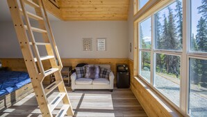 Interior acogedor de una caba ña con cama en altillo, sofá y amplios ventanales que dan a vistas de árboles.