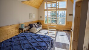 Un dormitorio con techo inclinado, paredes de madera, una cama con cubrecama azul, una escalera y un ventanal con vista a árboles.