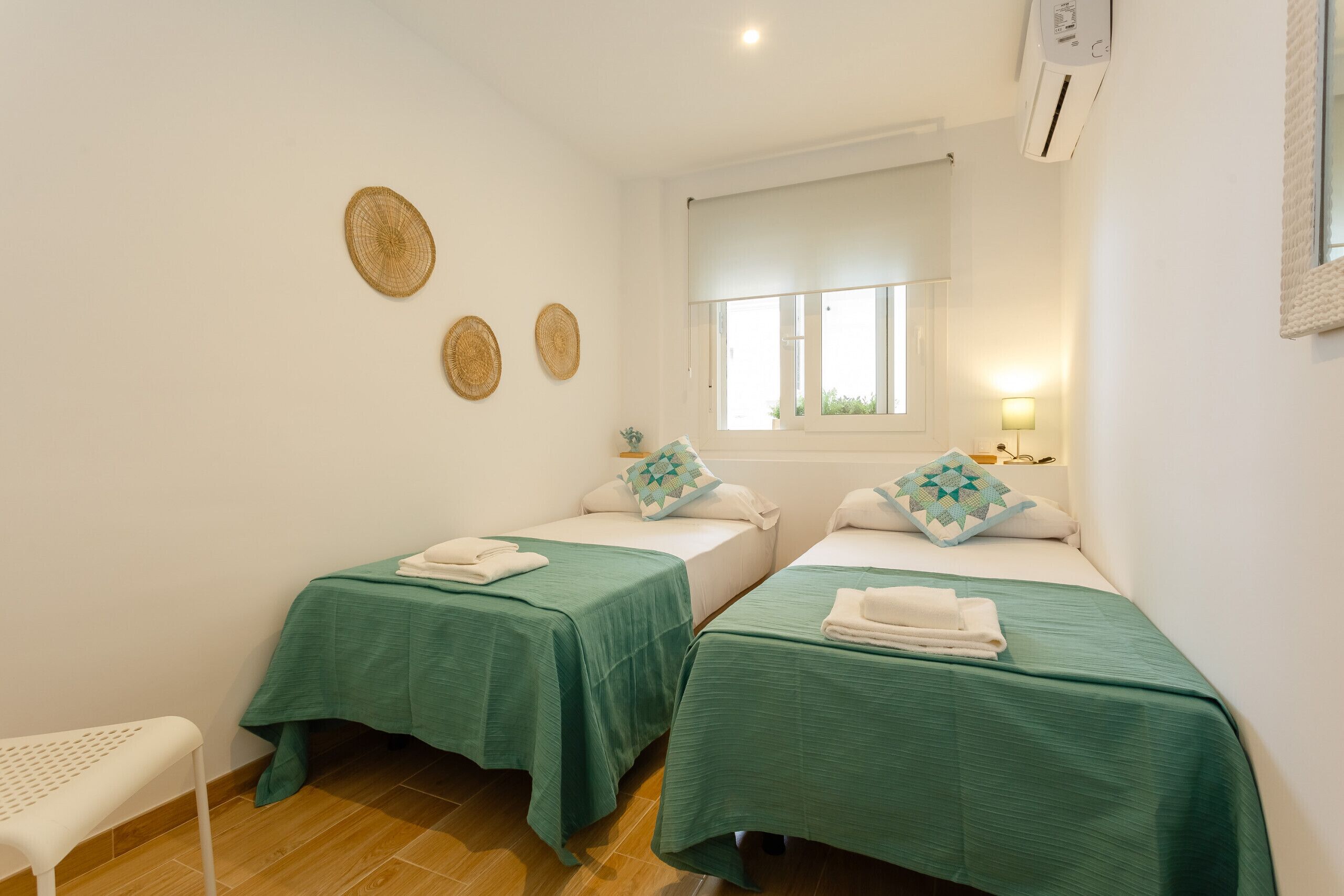 A room with two beds, each with a green bedspread and matching pillows, a small table with a lamp, and a chair.