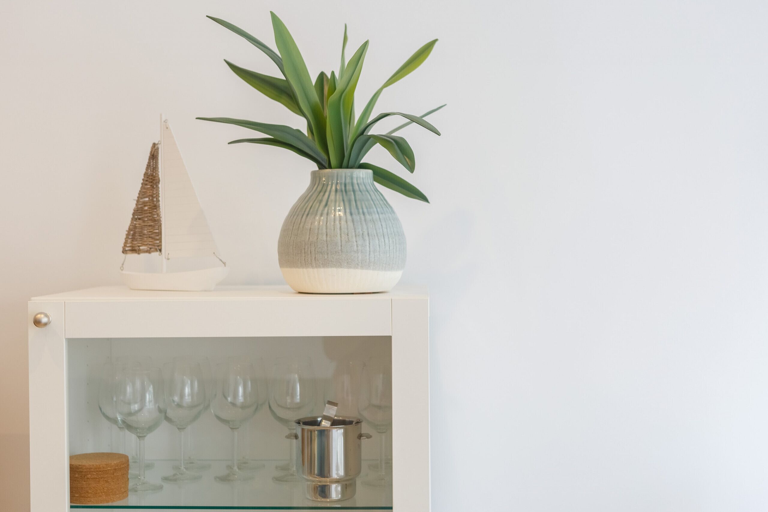 Un meuble blanc doté d’une vitrine où sont exposés des verres à vin et un pot en métal.