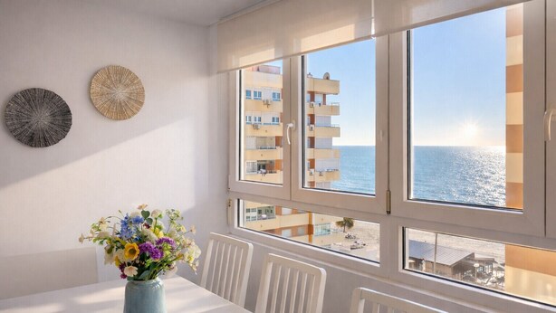 A room with a white table, a vase with flowers, and a view of the sea through large windows.