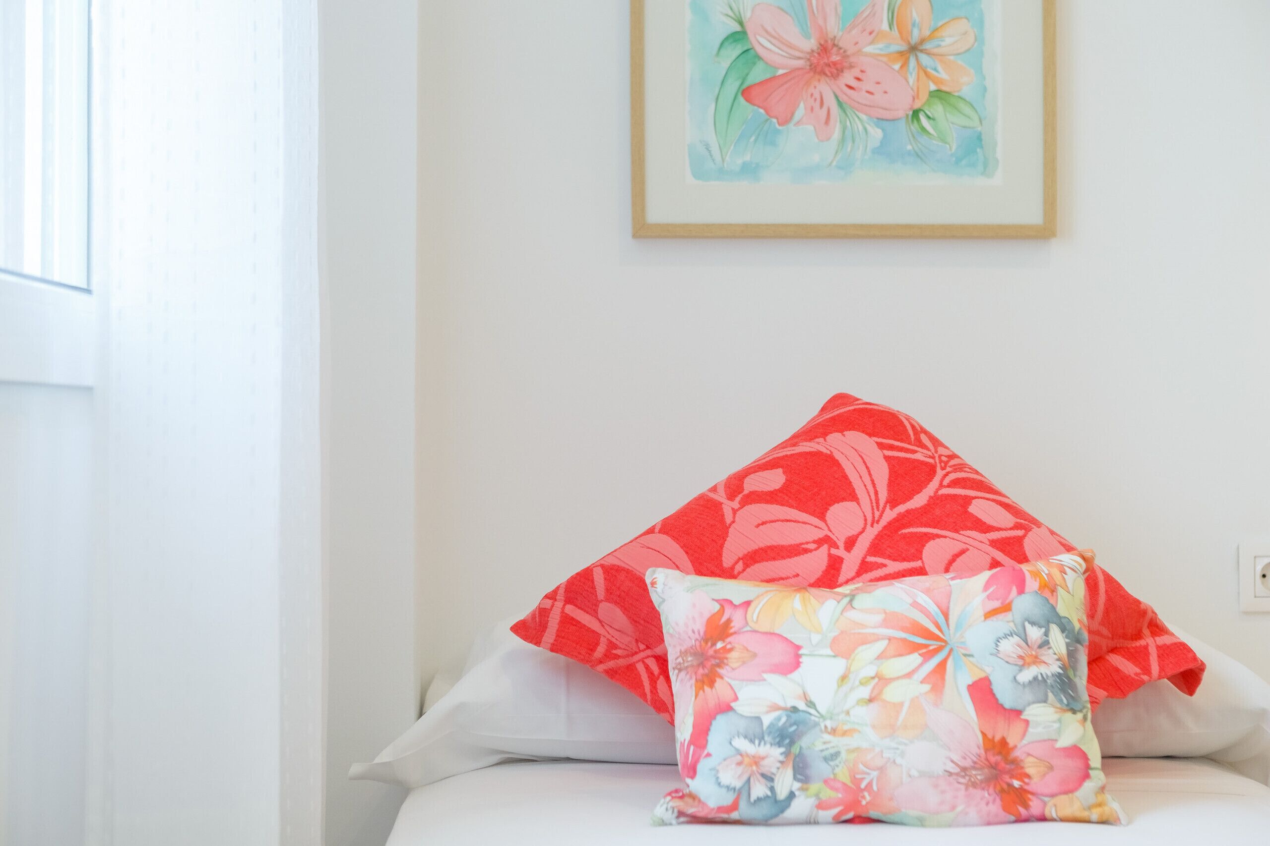 Una cama con ropa blanca y dos almohadas, una con estampado floral rojo y otra con estampado floral multicolores.